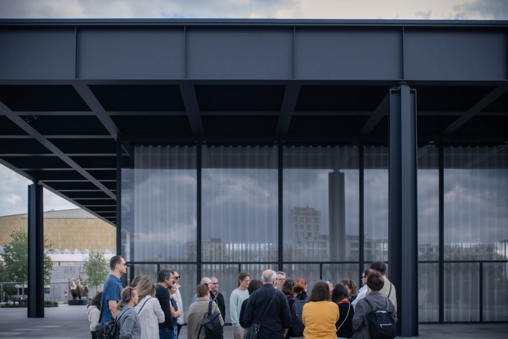 A group of people listening to a guide while standing next to tall glass facade of Mies van der Rohe's New national gallery. The massive horizontal slab is supported by columns around its perimeter.